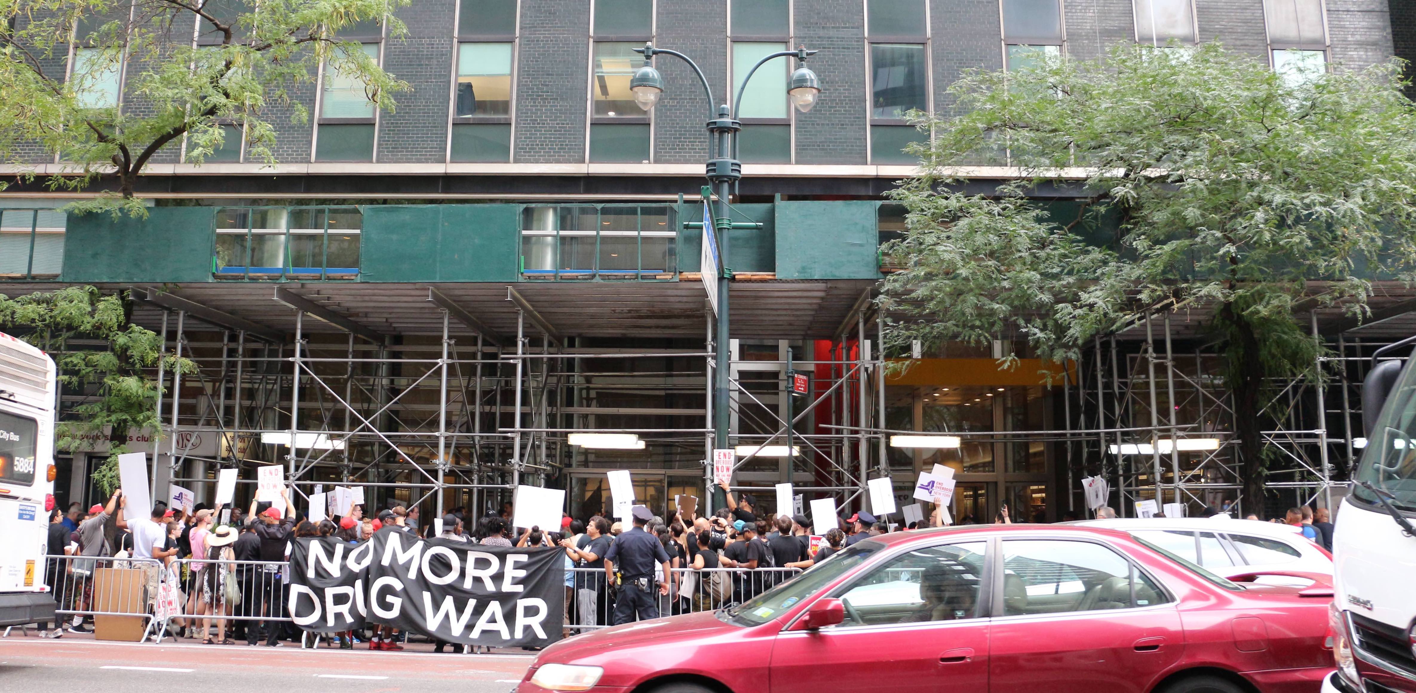 A group of protestors hold a banner outside a building. The banner reads 'NO MORE DRUG WAR'.