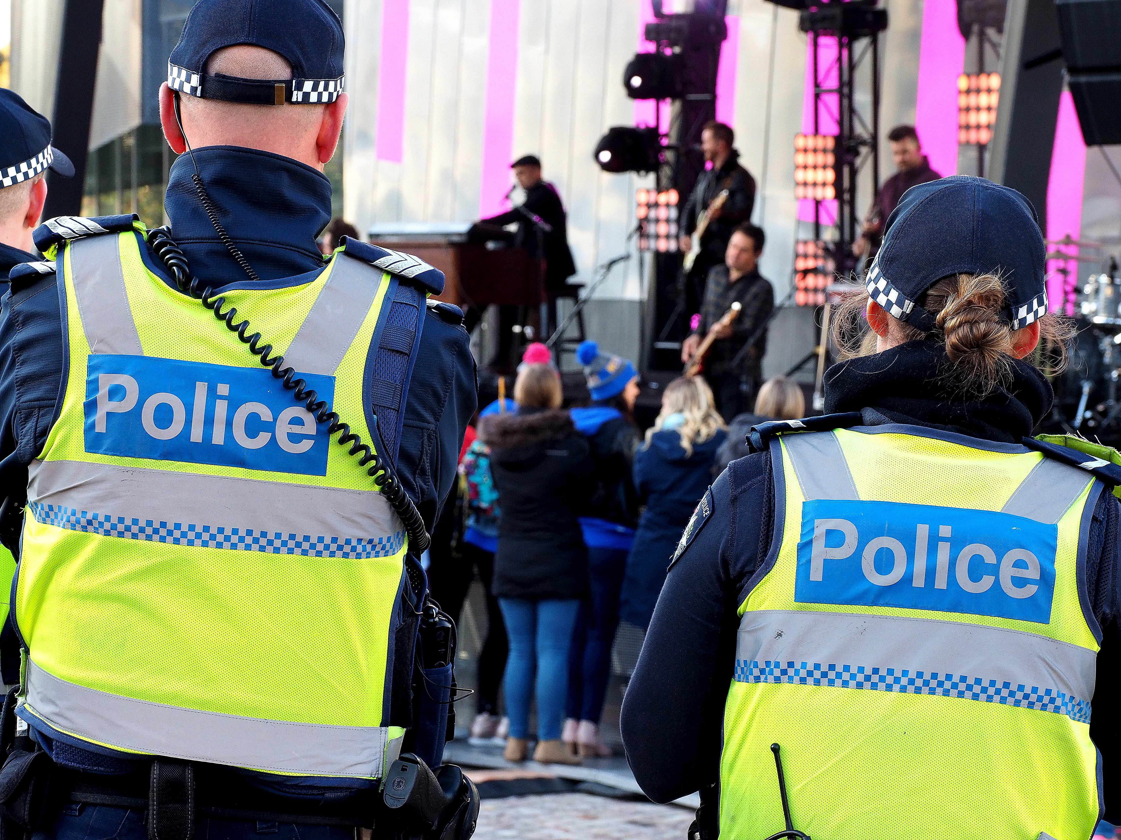 Two police people stand with their arms crossed, wearing yellow vests, observing a music concert.