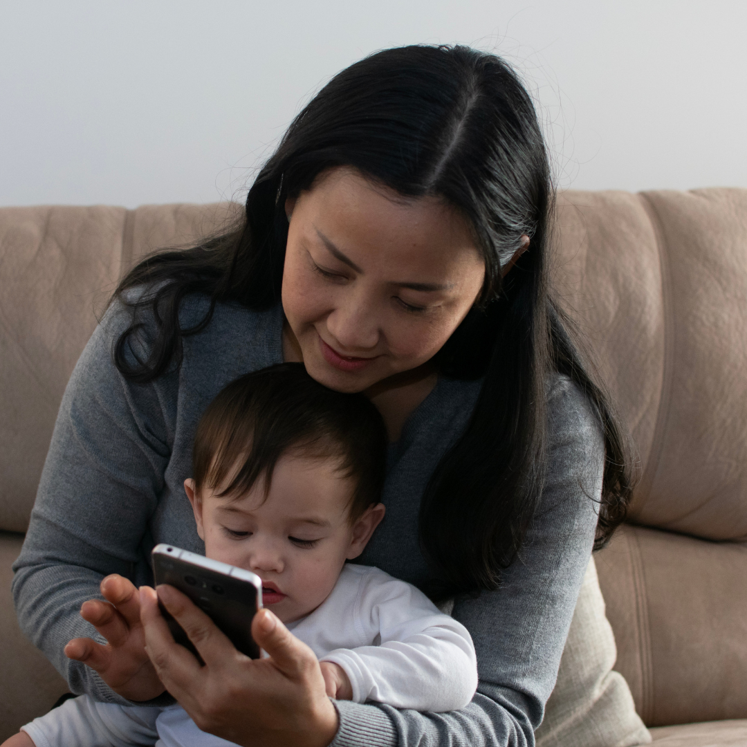 A woman in her thirties looks at her phone. She is sitting on a couch indoors with a young child. 