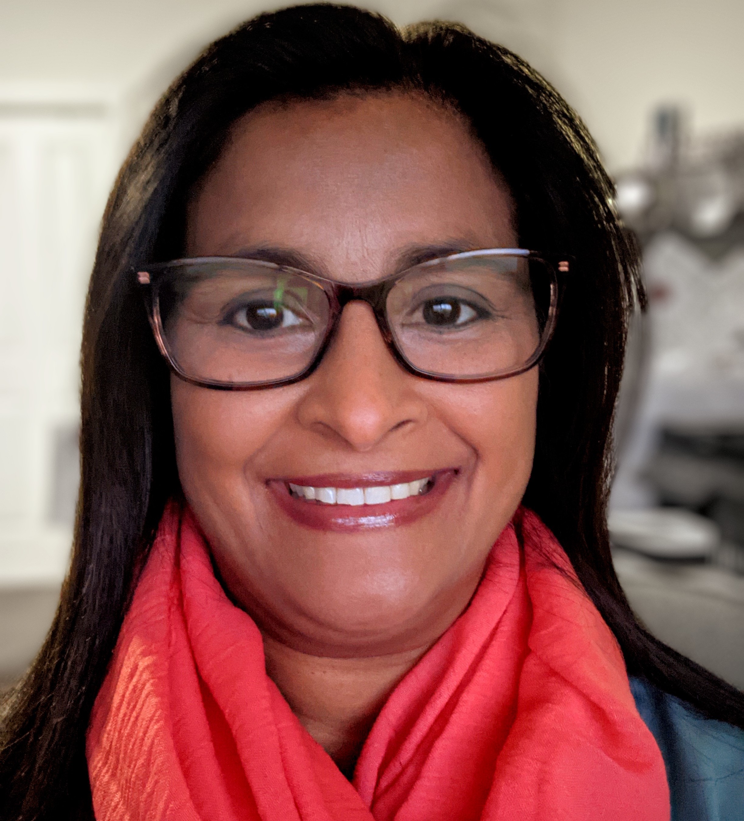 A photo of a woman smiling. She has brown skin, dark rimmed glasses and a red scarf.