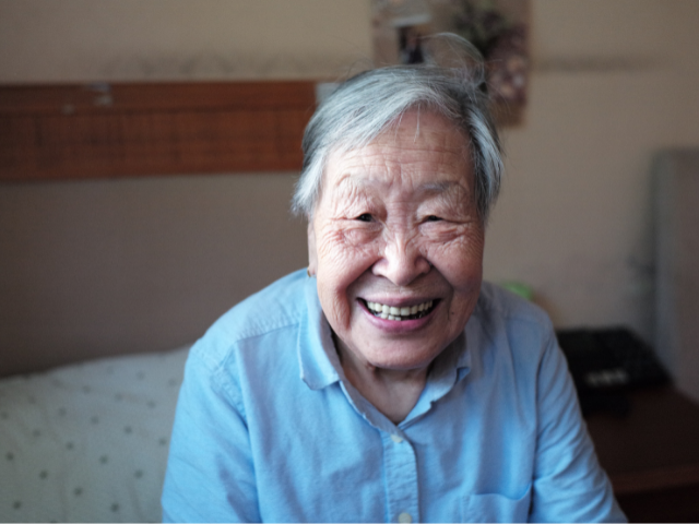 A photo of an older Chinese woman smiling at the camera. She is sitting on her bed and wearing a blue shirt.