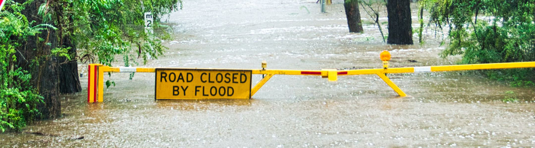 A yellow sign that reads 'Road closed by floods' over a flooded road.