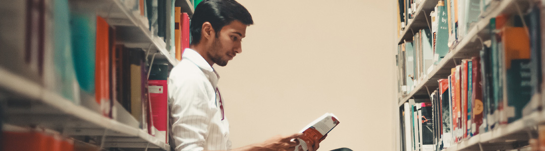 A young South Asian man sitting in the library and reading a book.