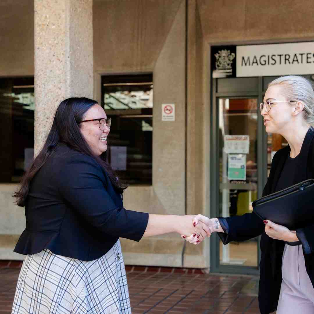 Two community legal centre workers smile and shake hands in front of a court house.