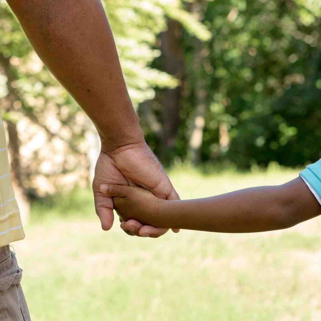 An adult and a child with brown skin hold hands.