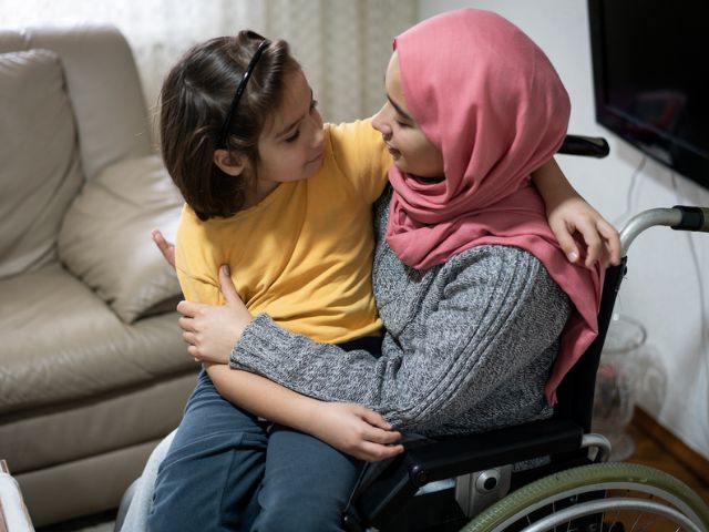 A woman and child smiling at each other. The woman is using a wheelchair and wears a pink headscarf; the child has short dark hair and is wearing a yellow t-shirt.