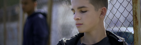 A young person looking down and leaning against a wire fence. There is another young person in the background.