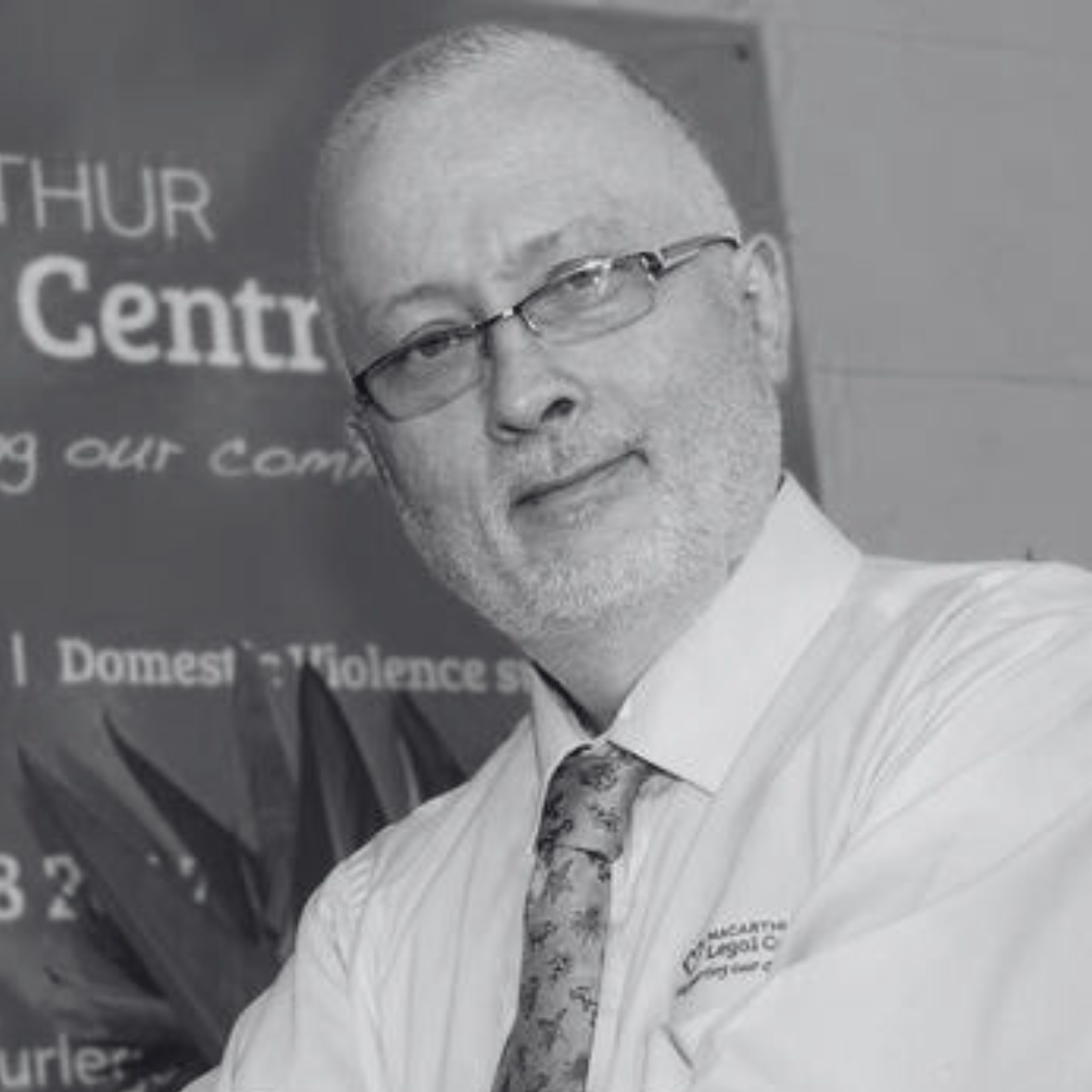 Robert smiling in front of a Macarthur Legal Centre sign. He wears glasses, a shirt and tie.