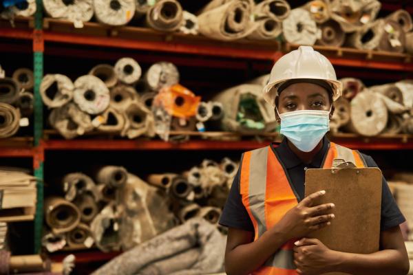 A young worker with dark skin in a workshop with rolls of fabric. She is wearing an orange high-vis vest, a face mask and a white hard hat.