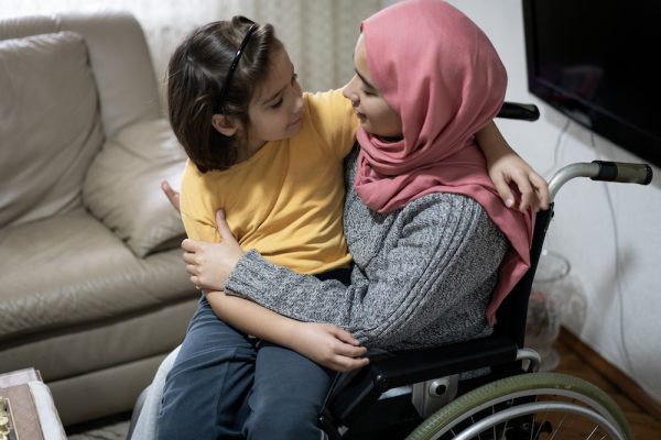 A woman with a pink head scarf sitting in a wheelchair hugs a young child. They are smiling at each other.